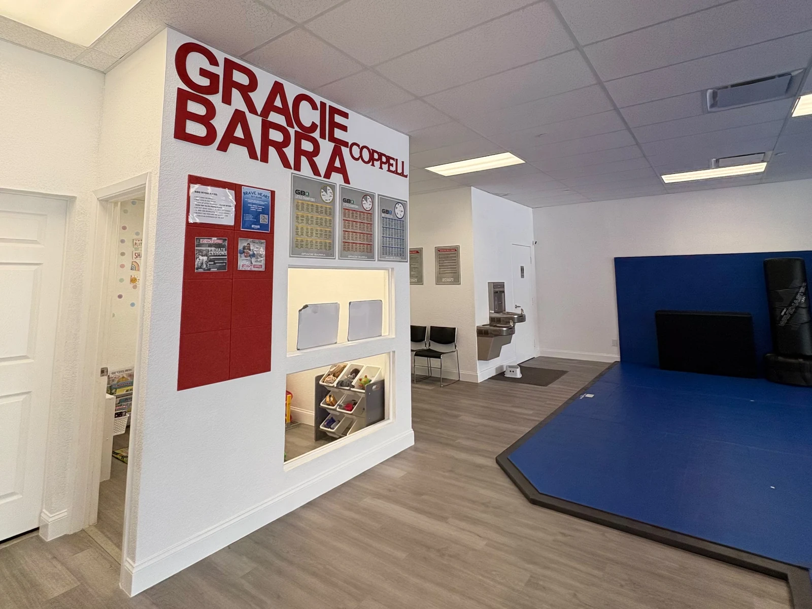 Front desk and lobby with the red Gracie Barra Coppell sign, schedule board, water fountain, and a Boxing-Thai mat area