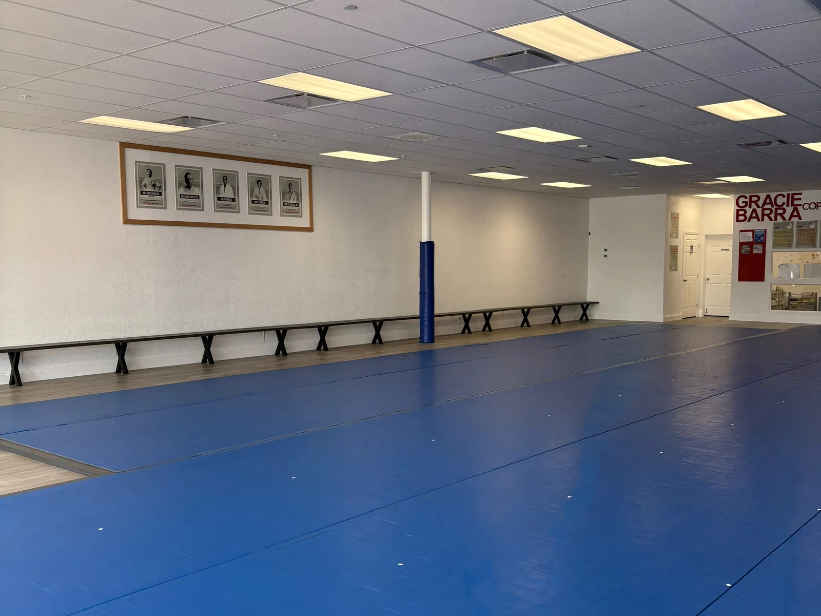 Wide view of the blue training mats at Gracie Barra Coppell, with benches along the wall and framed photos of the Gracie family